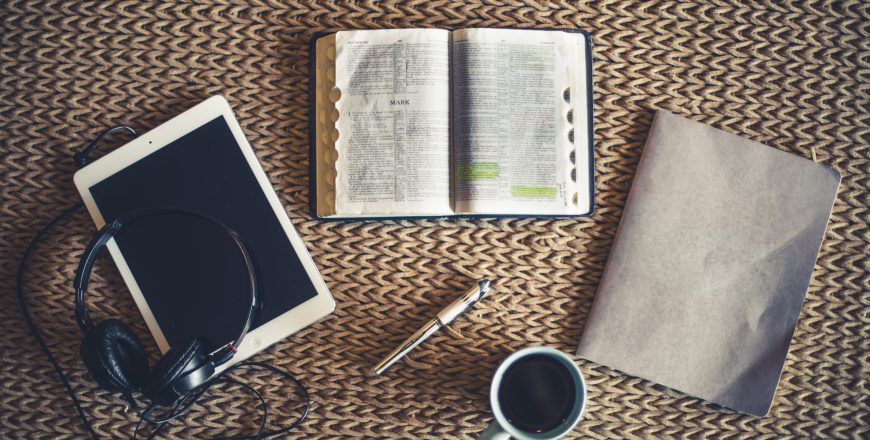 Shot from above of open Bible, coffee, notepad and tablet in fro